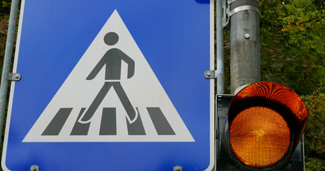 Ein Verkehrsschild mit Fußgängerüberweg und einer gelb leuchtenden Ampel signalisiert besondere Rücksichtnahme für den Fußverkehr im urbanen Raum.