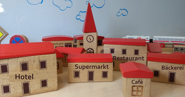 Mehrere Holzbausteine mit roten Dächern und Aufschriften wie „Hotel“, „Supermarkt“, „Restaurant“ und „Café“ stehen auf einem Tisch und bilden eine kleine Modellstadt ab.