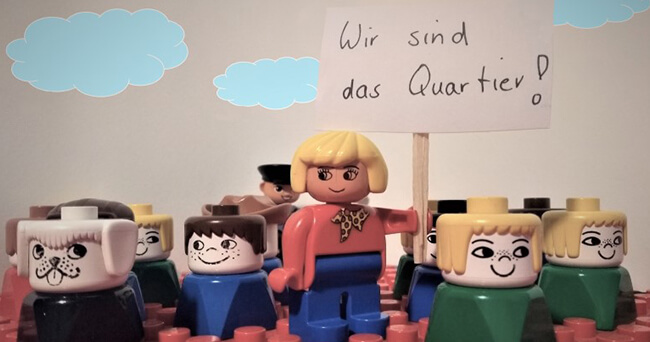 Mehrere Spielfiguren aus Kunststoff stehen auf roter Bauplatte, eine Figur hält ein Schild mit der Aufschrift „Wir sind das Quartier!“. Im Hintergrund weitere Figuren und gezeichnete Wolken.