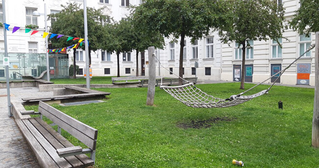 Begrünter Innenhof mit Hängematte, Holzbänken, Spiel- und Sitzflächen, im Hintergrund Wohngebäude und Bäume.