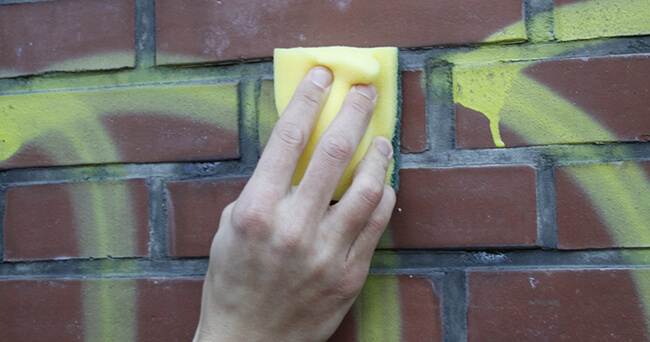 Eine Hand entfernt mit einem gelben Schwamm gelbe Graffiti-Farbe von einer roten Ziegelmauer.