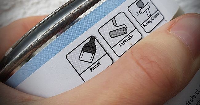 Nahaufnahme einer Hand, die eine Farbdose mit Symbolen für Pinsel, Lackrolle und Farbspritzgerät hält, geeignet für den Auftrag eines Graffitischutzanstrichs.