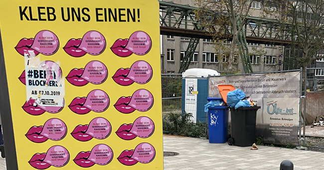 Eine gelbe Metalltafel mit zahlreichen pinken Stickern in Lippenform, auf denen steht „Kleb dich zur Sauberkeit“. Daneben ein Bauzaun, Mülltonnen und eine Baustelle im Hintergrund. Die Tafel dient zur Sammlung von Kaugummis im öffentlichen Raum und wird „Gum Wall“ genannt.