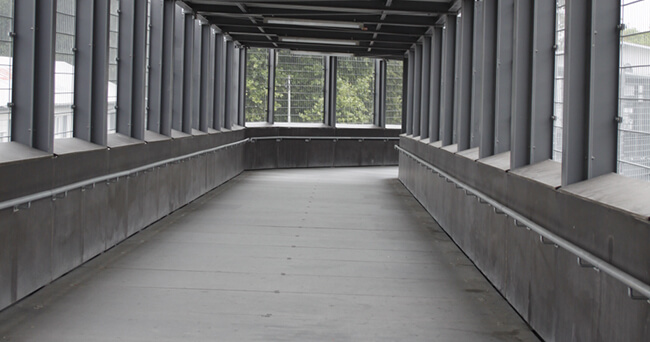 Überdachte, gut einsehbare Fußgängerbrücke mit freier Sichtachse und seitlichen Fenstern, die Übersichtlichkeit und soziale Kontrolle im öffentlichen Raum symbolisiert.