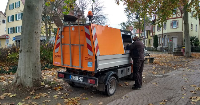 Ein Mitarbeiter der Stadtreinigung steht neben einem orangefarbenen Reinigungsfahrzeug in einer von Bäumen gesäumten Wohnstraße, Herbstlaub liegt auf dem Boden.