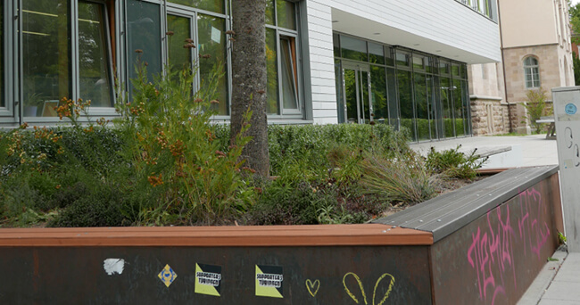 Ein bepflanztes Hochbeet mit Sitzfläche vor einem modernen öffentlichen Gebäude, an der Außenseite des Beetes sind Sticker und Graffiti sichtbar.