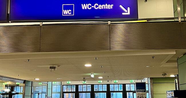 Innenaufnahme eines öffentlichen Gebäudes mit einem blauen Hinweisschild, auf dem WC-Center und einem Pfeil nach rechts, steht. Darunter sind Türen und Informationsschilder zu sehen.