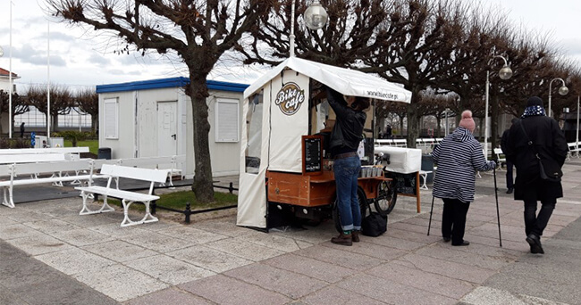 Ein mobiler Kaffeestand in einem weißen Zeltaufbau steht auf einem gepflasterten Platz. Eine Person bedient den Stand, während drei ältere Menschen davorstehen. Im Hintergrund kahle Bäume, Bänke und ein Toilettenhäuschen.