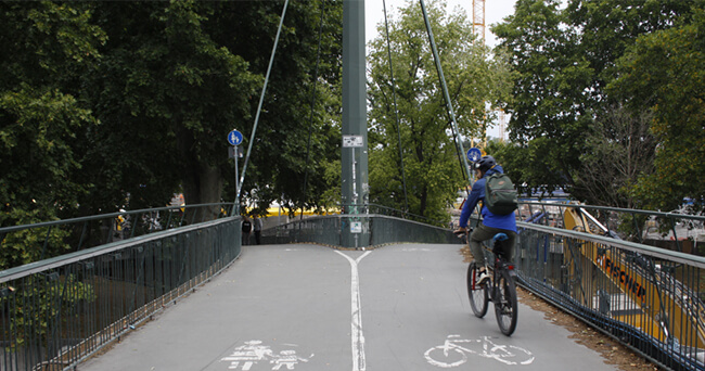 Ein Fuß- und Radweg auf einer Brücke ist durch eine weiße Linie in zwei Bereiche unterteilt. Auf dem linken Bereich ist ein Fußgängersymbol, auf dem rechten ein Fahrradsymbol aufgebracht. Ein Radfahrer nutzt die rechte Spur. Im Hintergrund sind Bäume und weitere Personen erkennbar.