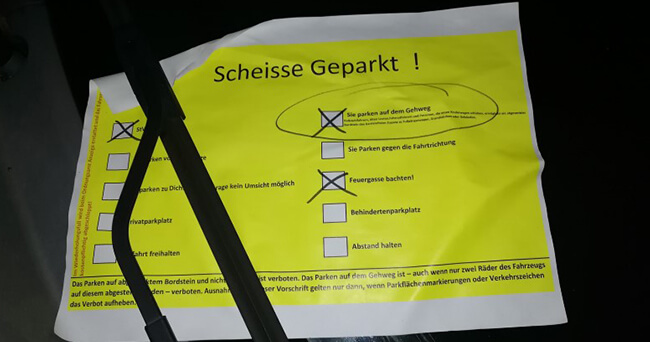 Ein gelber Hinweiszettel mit dem Titel „Scheisse geparkt!“ liegt unter einem Scheibenwischer. Der Zettel listet verschiedene Parkverstöße auf, wie "„Sie parken auf dem Gehweg“ oder „Feuergasse blockiert“, und ist teils angekreuzt und markiert.