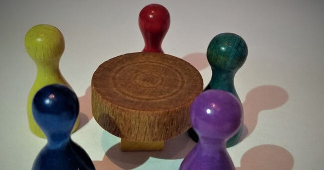 Nahaufnahme von fünf unterschiedlich farbigen Spielfiguren, die um einen kleinen, runden Holztisch angeordnet sind.