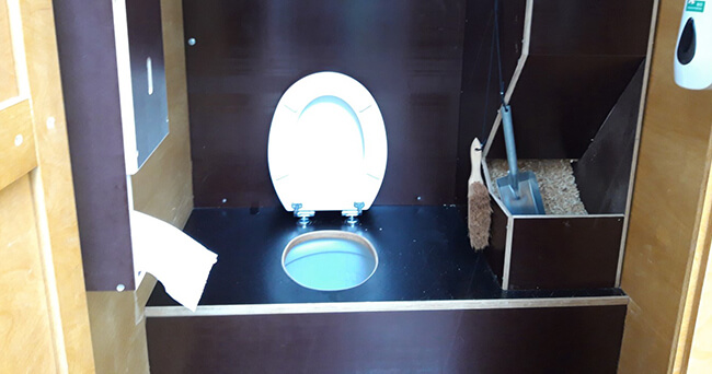 Innenraum einer mobilen Komposttoilette aus Holz mit geöffneter Toilettenbrille, Toilettenpapier, Bürste und Fach mit Einstreu.