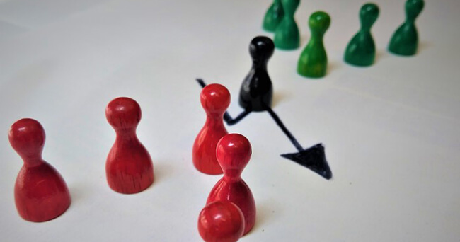 Symbolbild mit bunten Spielfiguren auf einem weißen Untergrund. Eine schwarze Figur vermittelt zwischen zwei Gruppen aus roten und grünen Figuren – dargestellt durch einen Pfeil.