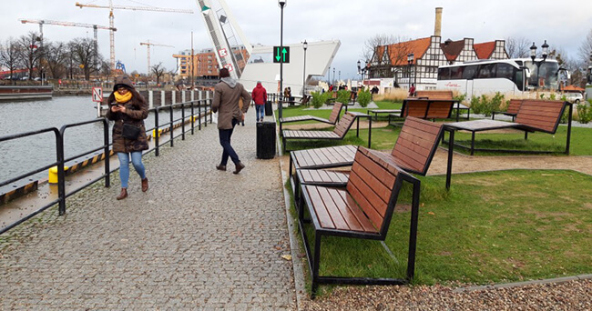 Mehrere Holzsitzbänke und Liegen stehen auf einer Grünfläche an einer gepflasterten Promenade am Wasser; Passanten gehen vorbei, im Hintergrund sind Baukräne und Gebäude zu sehen.