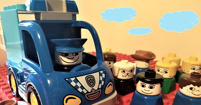 Mehrere Spielfiguren, darunter eine Polizeifigur in einem blauen Spielzeug-Polizeifahrzeug, stehen vor einer Gruppe weiterer Figuren auf roter Noppenplatte mit eingezeichneten Wolken im Hintergrund.