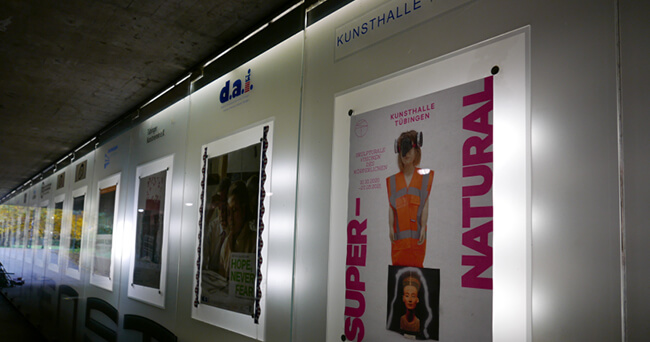 Seitliche Ansicht einer beleuchteten Plakatwand der Kunsthalle in einem Tunnel, mit verschiedenen Postern zu aktuellen und vergangenen Kulturveranstaltungen.