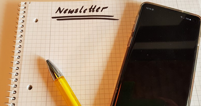 Auf einem karierten Notizblock liegt ein gelber Kugelschreiber, darüber steht handschriftlich das Wort „Newsletter“. Rechts daneben liegt ein Smartphone mit schwarzem Display.