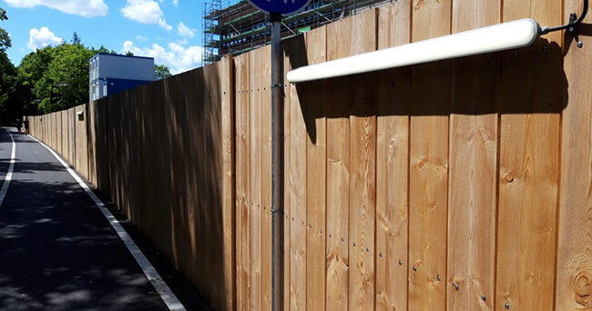 Eine hohe Lärmschutzwand aus senkrechten Holzelementen steht entlang eines asphaltierten Wegs, der an einer Baustelle vorbeiführt. Davor verläuft ein kombinierter Rad- und Fußweg unter blauem Himmel.