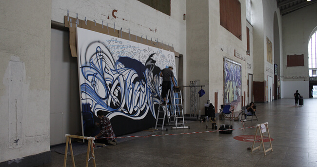 Mehrere Personen gestalten in einer großen Halle eine legale Graffiti-Fläche mit bunten Sprayfarben, eine Leiter und Malutensilien stehen bereit.