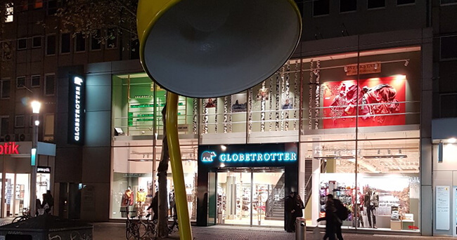 Eine übergroße gelbe Straßenlampe im Vordergrund, dahinter ein hell erleuchtetes Geschäft mit Glasfassade in einer Innenstadt bei Nacht.