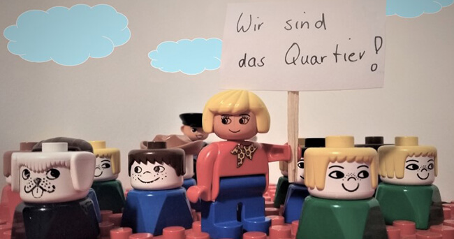 Mehrere bunte Spielzeugfiguren stehen dicht beisammen, eine Figur in der Mitte hält ein Schild mit der Aufschrift „Wir sind das Quartier!“