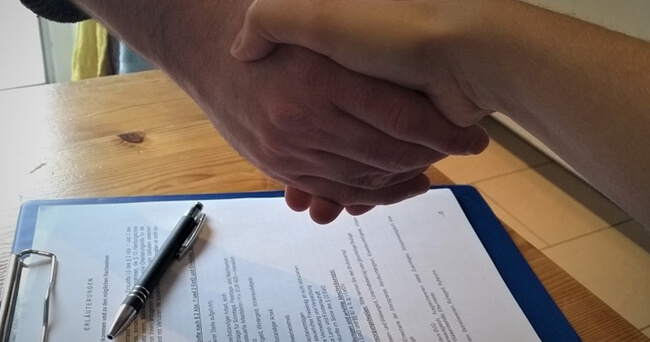 Zwei Menschen geben sich zur Vertragsunterzeichnung die Hand. Im Vordergrund liegt ein Klemmbrett mit einem unterschriftsbereiten Formular und einem Kugelschreiber auf einem Holztisch.