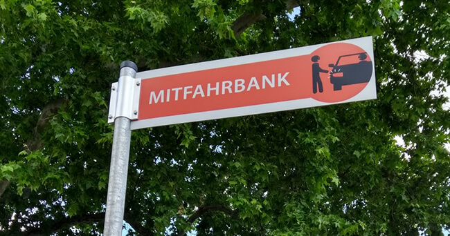 Straßenschild mit der Aufschrift „Mitfahrbank“ und einer Piktogramm-Darstellung einer Person, die zu einem Auto geht, im Hintergrund ein grüner Laubbaum.