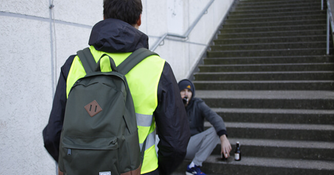 Eine Person mit Warnweste steht vor einer sitzenden Person auf einer Treppe im öffentlichen Raum. Die stehende Person trägt einen Rucksack und spricht offenbar mit der anderen Person, die eine Bierflasche neben sich stehen hat.