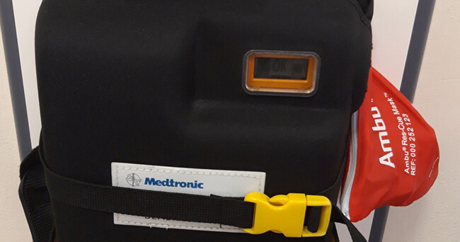 Eine schwarze Tragetasche mit Medizintechnik-Aufdruck, seitlich befestigter roter Beatmungsmaske und gelbem Verschluss, bereit für den Einsatz in der mobilen ärztlichen Versorgung.