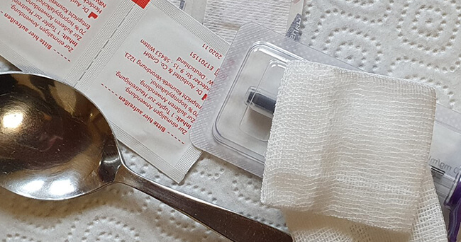 Verschiedene sterile Utensilien zur sicheren Durchführung eines Drogenkonsums, darunter ein Löffel, Alkoholtupfer, Kanüle und Mullkompresse, ausgelegt auf einer Papierserviette.