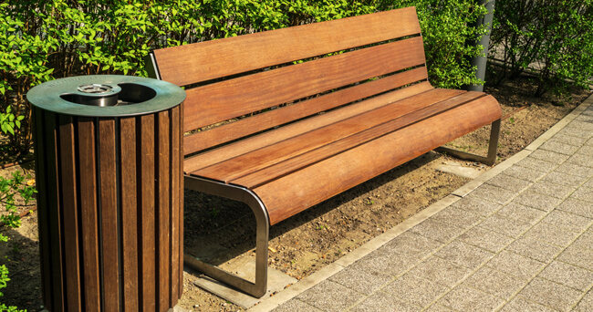 Ein moderner, brauner Mülleimer aus Holz steht neben einer passenden Sitzbank auf einem gepflasterten Gehweg, eingerahmt von grünen Hecken.