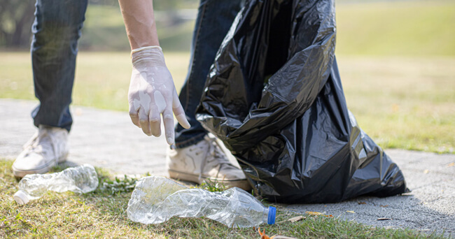 Eine Person in Turnschuhen und mit Einweghandschuhen hebt eine leere Plastikflasche vom Boden auf und steckt sie in einen schwarzen Müllsack, umgeben von Rasenfläche und Gehweg.