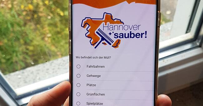 Hand hält Smartphone mit geöffneter Müllmelde-App „Hannover sauber!“ vor einem Fenster, Auswahlmöglichkeiten zur Lage des Mülls sichtbar.
