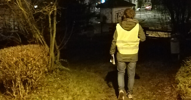 Eine Person mit gelber Warnweste geht nachts auf einem beleuchteten Weg in einem Park. Im Hintergrund sind Büsche, Bäume und eine leicht beleuchtete Straße zu sehen.