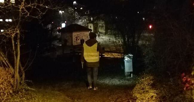 Eine Person mit Warnweste geht in der Dunkelheit auf einem unbeleuchteten Weg in Richtung Stadtgebiet, begleitet von schwacher Straßenbeleuchtung im Hintergrund.