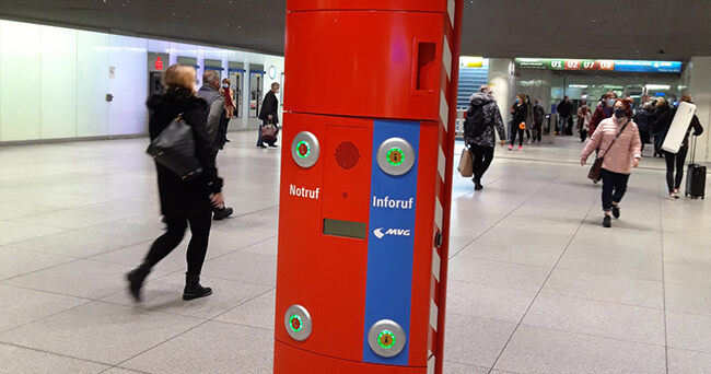 Rote Notrufsäule mit Notruf- und Inforufknöpfen in einer belebten Bahnhofshalle, Menschen gehen im Hintergrund vorbei.