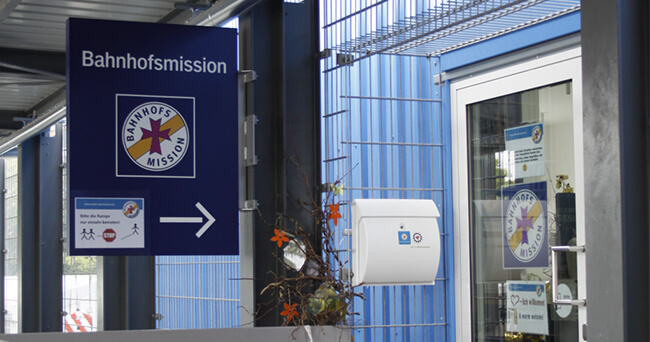Blaues Schild mit der Aufschrift „Bahnhofsmission“ und Pfeil weist auf den Eingang einer Einrichtung hin, die Notschlafplätze für wohnungslose Menschen anbietet. Im Hintergrund Glasfassade mit Tür und weiteren Beschilderungen.