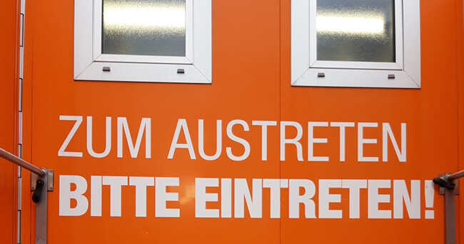 Orangefarbener Container mit zwei Fenstern und großflächiger weißer Aufschrift „Zum Austreten bitte eintreten!“, aufgenommen von vorne.