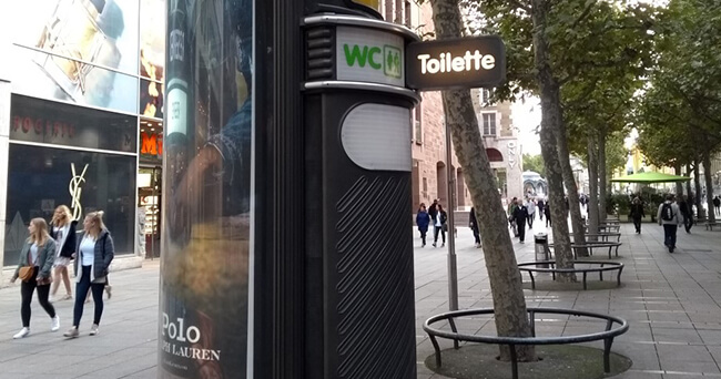 Öffentliche Toilettenanlage an einer Einkaufsstraße mit beleuchtetem WC-Schild und Bäumen im Hintergrund.