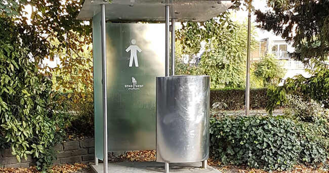 Ein modernes öffentliches Urinal mit Sichtschutz und Edelstahlwand steht in einer von Bäumen und Büschen umgebenen Grünanlage. Auf dem Sichtschutz ist ein Piktogramm einer männlichen Figur zu sehen.