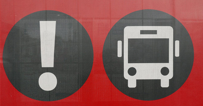 Zwei weiße Piktogramme auf schwarzem Kreis vor rotem Hintergrund: links ein Ausrufezeichen, recht ein stilisierter Linienbus, symbolisch für Aufmerksamkeit im öffentlichen Nahverkehr.