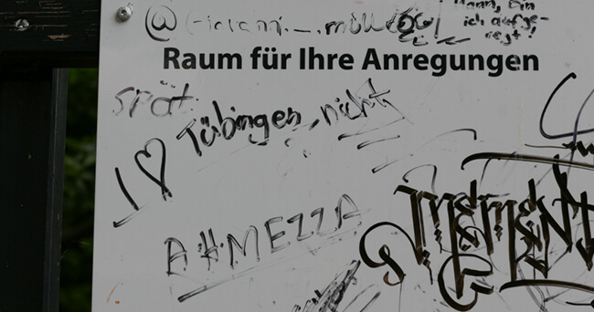 Ein weißes Hinweisschild mit der Aufschrift „Raum für Ihre Anregungen“ ist mit Kritzeleien, Graffiti und teils lesbaren Kommentaren wie „ich liebe Tübingen nicht“ beschmiert.