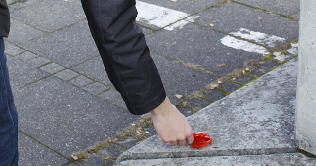Eine Person im dunklen Mantel beugt sich vor und hebt ein rotes Stück Plastikmüll von einer Betonfläche an einer Straßenkreuzung auf.