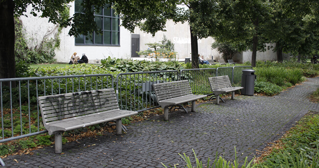 Drei Holzbänke stehen entlang eines gepflasterten Weges in einer begrünten Parkanlage. Im Hintergrund sitzen Menschen auf einer Rasenfläche neben einem Gebäude.