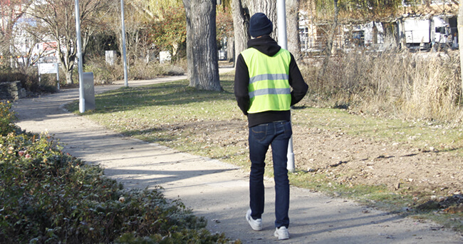 Eine Person in Jeans, Kapuzenjacke und gelber Warnweste geht einen geteerten, sandigen weg in einem Park entlang. Umgeben ist die Szene von Bäumen, Büschen und Wiesen.