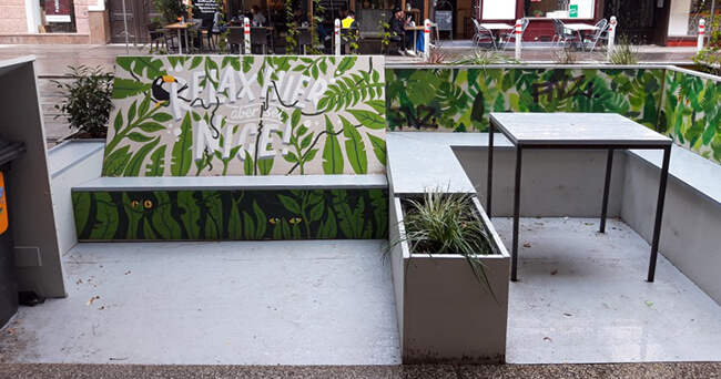 Ein aufwendig bemaltes Parklet mit grünem Blättermuster bietet Sitzgelegenheiten und einen Tisch auf einer umgenutzten Fläche am Straßenrand. Auf der Rückenlehne steht „Relax, aber safe“.