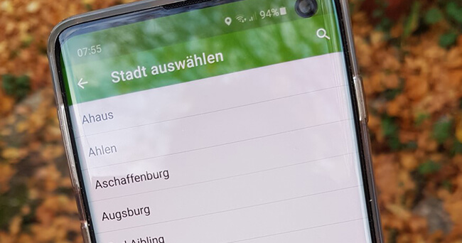 Ein Smartphone zeigt eine geöffnete App mit der Überschrift „Stadt auswählen“. Auf dem Display sind Städtenamen wie Ahaus, Ahlen und Aschaffenburg sichtbar. Im Hintergrund liegt herbstlicher Laub.