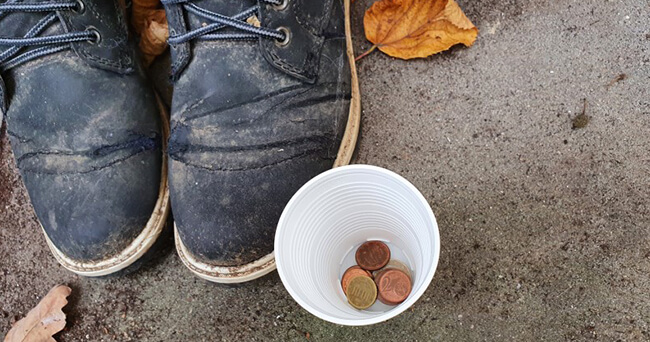 Ein Paar stark abgetragene schwarze Schuhe steht auf einem Gehweg neben einem weißen Plastikbecher, in dem sich einige Cent-Münzen befinden. Daneben liegen trockene Herbstblätter.