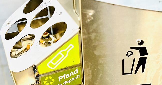 Ein silberner Mülleimer mit aufgesetztem Pfandflaschenhalter aus Metall zeigt ein Symbol zum Einwerfen von Flaschen sowie das Wort „Pfand“ in Deutsch und Englisch.