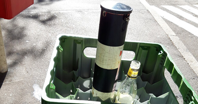 Eine grüne Getränkekiste mit leeren Glasflaschen steht auf dem Gehweg. In der Mitte der Kiste ist ein schwarz-weißer Poller platziert. Im Hintergrund ist ein Sperrfläche zu erkennen.